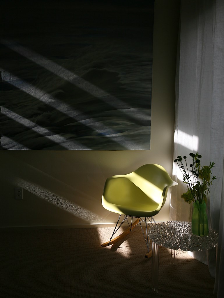 Dark cozy room with beige walls and green chair with silver side table and green vase and large beige and blue painting