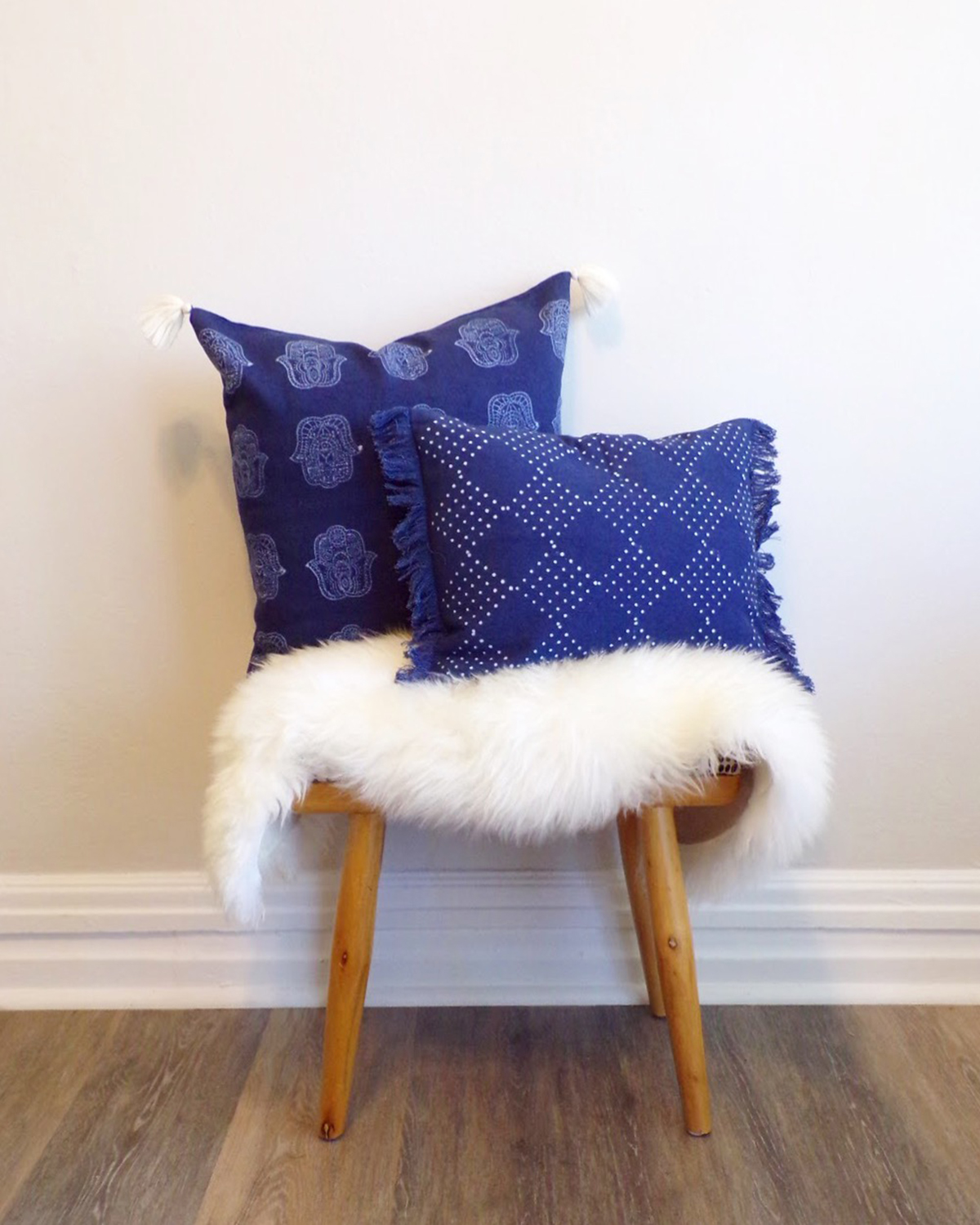 Shot of two pillows with blue pillow cases sitting on a throw and stool. Pillows are covered with Chelsea's design