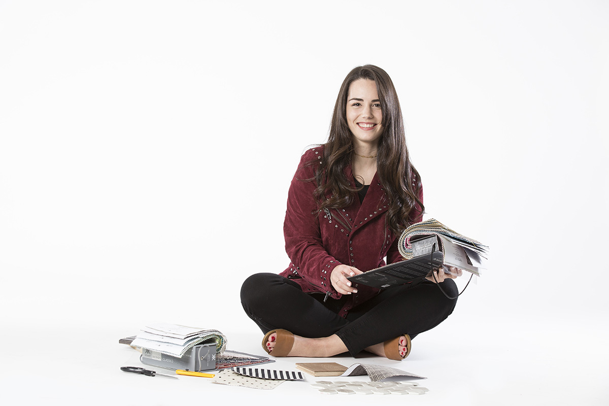 Photo of DI student Britnie Cowling sitting on the floor, looking at fabric samples, smiling at the camera