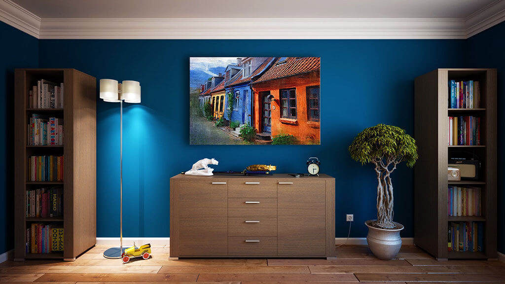 Interior Design Trends 2020 - Bold Colors. A stylized shot of an interior space, showing royal blue painted walls, with a brown credenza in the center. There is a photo of houses hanging above the credenza