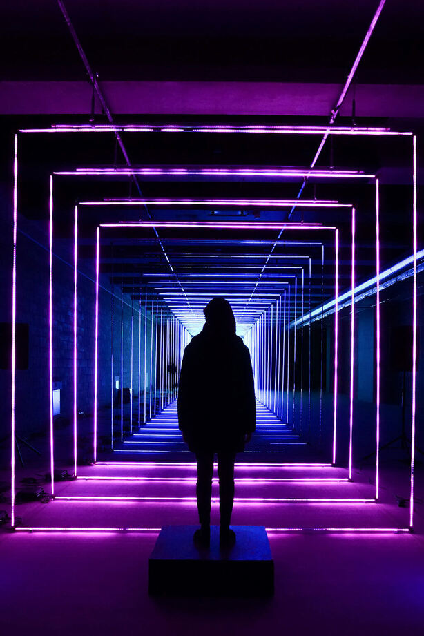 Escapism. There is a person standing at the end of a hallway with fluorescent purple neon lights shaped as a squares and framing the hallway