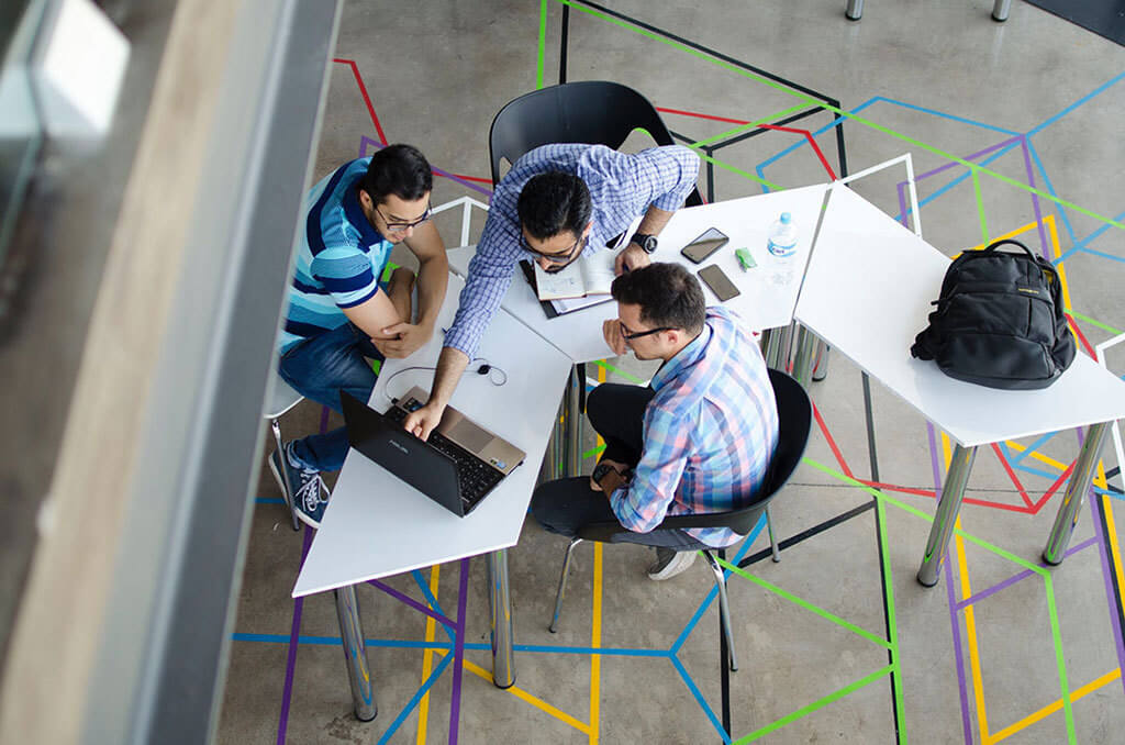 Interior Design Trends 2020 - Shift from Private to Shared Space. A bird's eye view of three people huddled at a table, looking at a laptop.